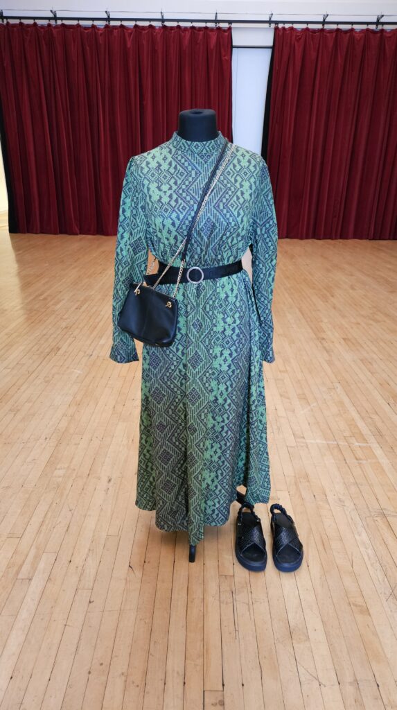 A mannequin dressed in a green patterned maxi dress with a belt and cross-body bag, with black sandals beside it
