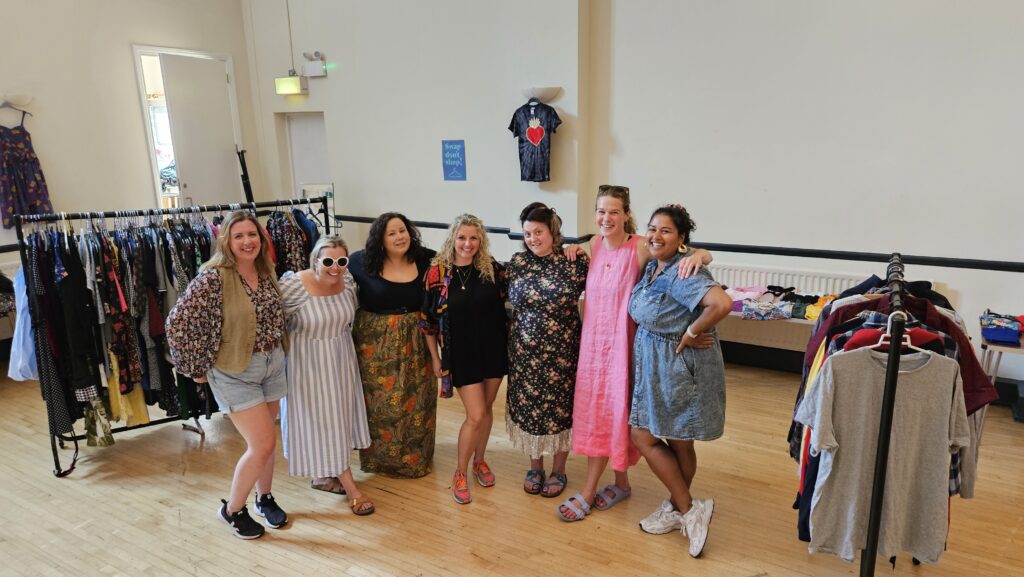 Seven smiling swappers with their arms around each other in a hall lined with clothes rails