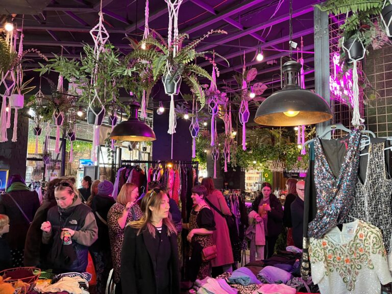 Swappers browsing rails of clothes under hanging plants and purple lights at an indoor swap